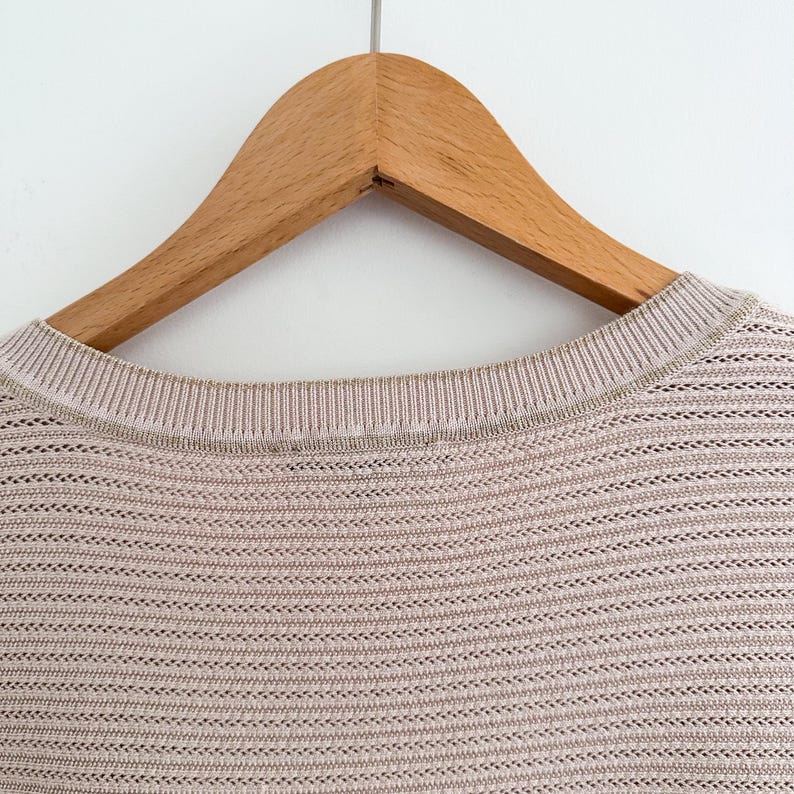 May include: Close-up of a light brown knit sweater with a round neckline. The sweater is hanging on a wooden hanger against a white background. The knit pattern is horizontal with small openings throughout the fabric.