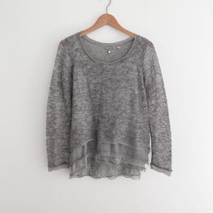 May include: A gray long-sleeved sweater with a scoop neck and a layered, ruffled hem. The sweater is hanging on a wooden hanger against a white background. The fabric appears to be a soft, textured knit.