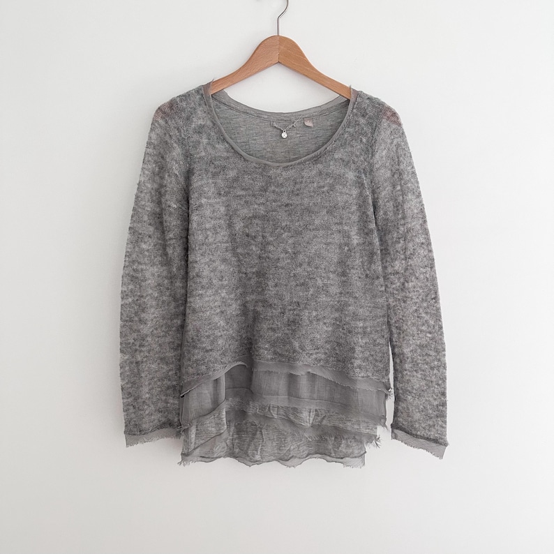May include: A gray long-sleeved sweater with a scoop neck and a layered, ruffled hem. The sweater is hanging on a wooden hanger against a white background. The fabric appears to be a soft, textured knit.