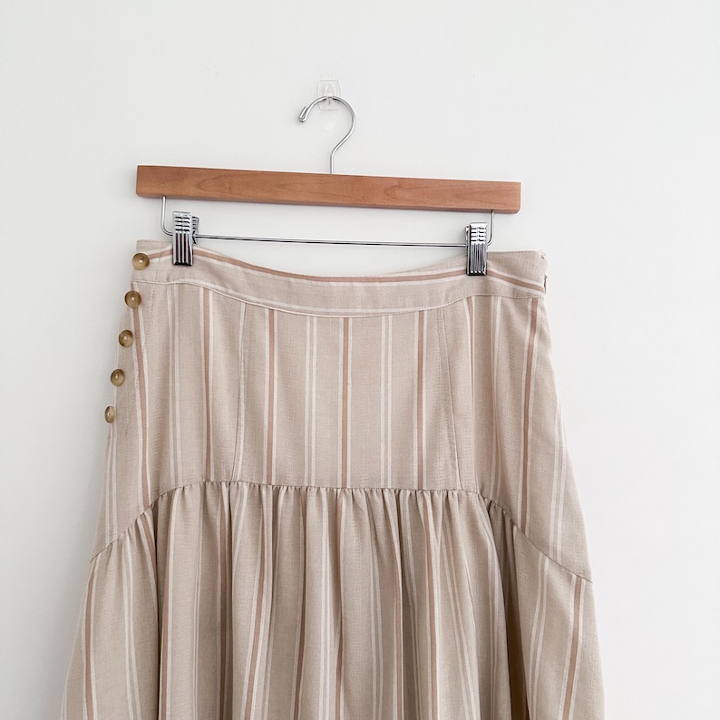 May include: A beige and white striped skirt with a button detail on the side. The skirt is hanging on a wooden hanger against a white background. The skirt has a high waist and a flared hem.