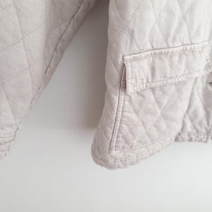 May include: A light beige quilted jacket with a diamond pattern. The jacket has a pocket on the front and a button closure. The fabric appears to be a soft, lightweight material. The jacket is shown against a white background.