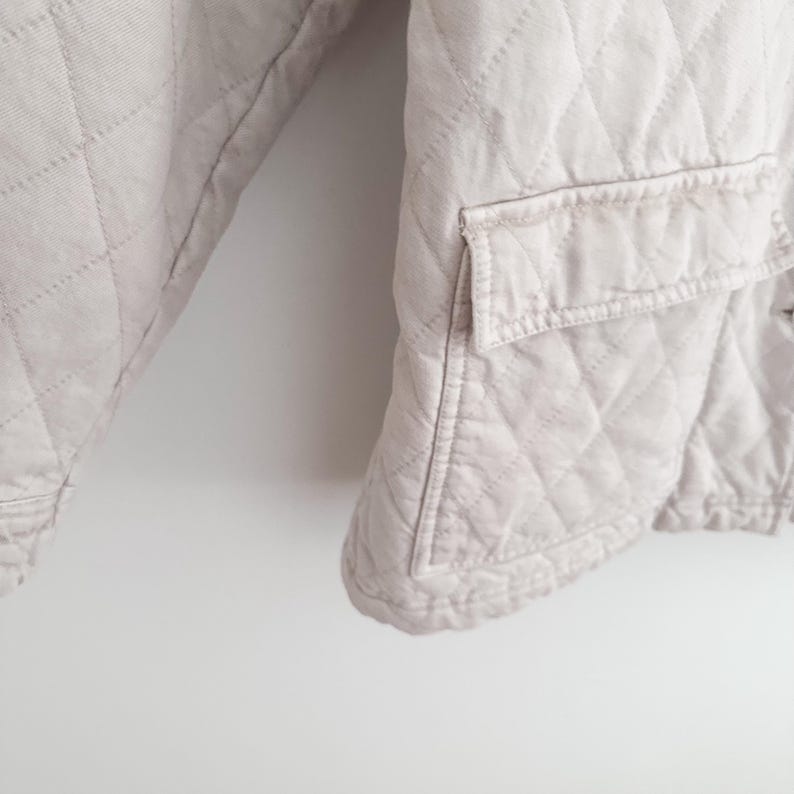 May include: A light beige quilted jacket with a diamond pattern. The jacket has a pocket on the front and a button closure. The fabric appears to be a soft, lightweight material. The jacket is shown against a white background.
