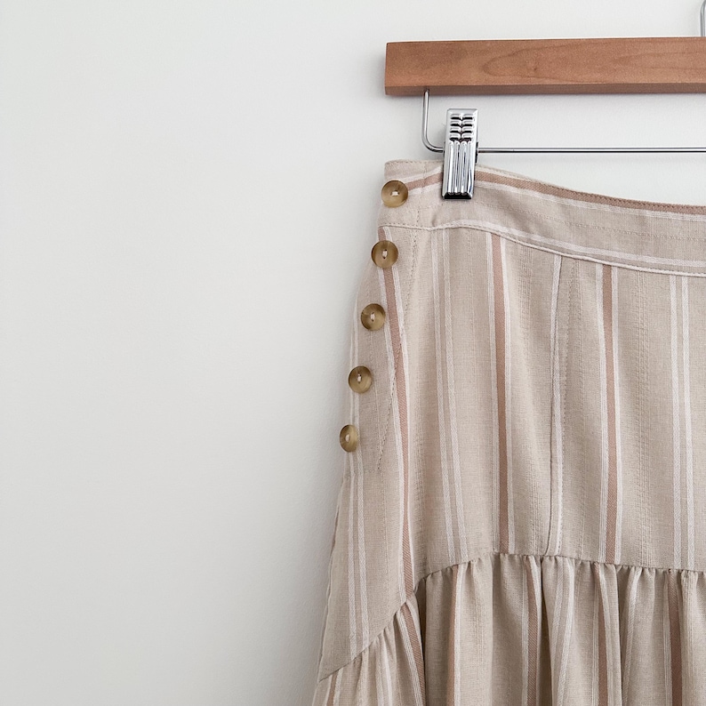 May include: A beige and white striped skirt with a button-up side detail. The skirt is hanging on a wooden hanger. The buttons are a light brown color. The skirt has a tiered, ruffled hemline.