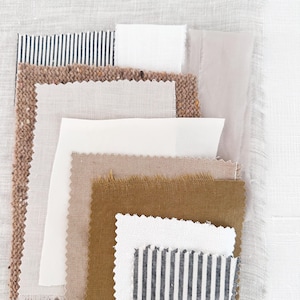 May include: A collection of fabric swatches in various textures and colors. The swatches include white, beige, brown, and striped patterns. Some have frayed edges, while others have clean cuts. The fabrics are arranged on a light-colored background.