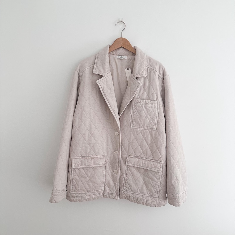 May include: A light beige quilted jacket with a classic collar and two front pockets. The jacket is hanging on a wooden hanger against a white background. The jacket has a diamond pattern.