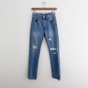 May include: Light blue denim jeans with distressed details, including rips and fading. The jeans are hung on a wooden hanger against a white background. The jeans have a button fly and classic five-pocket design.