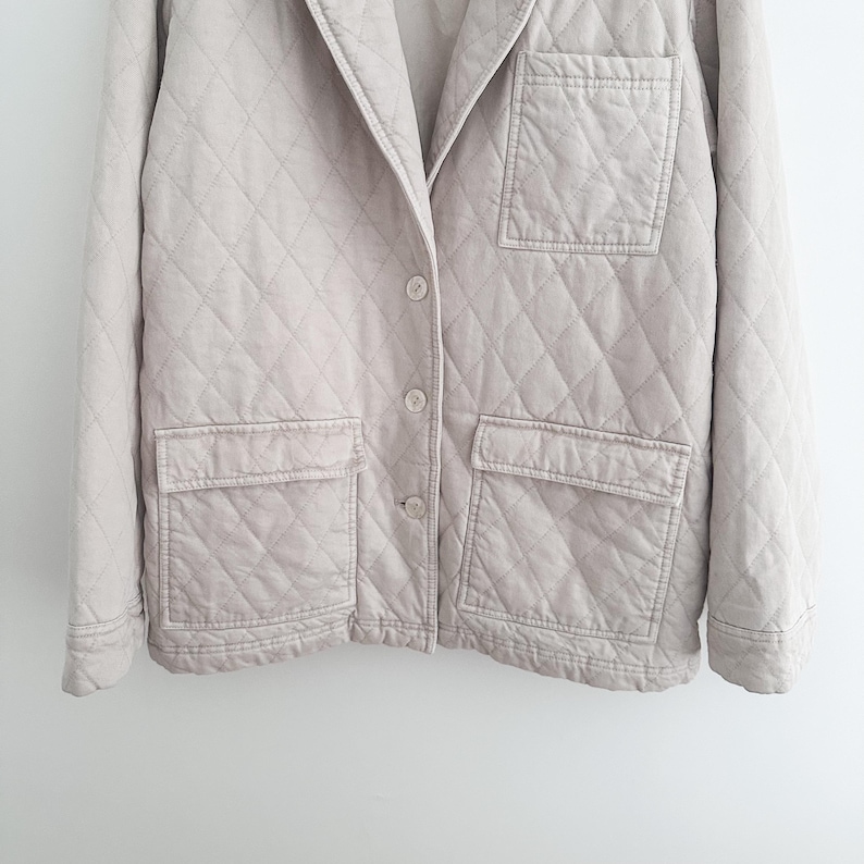 May include: A light beige quilted jacket with a notched collar, three buttons, and two front pockets. The jacket has a diamond pattern throughout and is made of a soft, textured fabric.