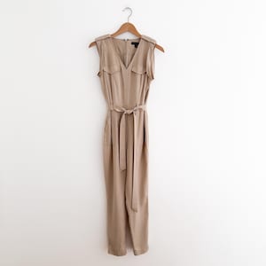 May include: A sleeveless tan jumpsuit with a V-neck, two small chest pockets, and a matching fabric belt. The jumpsuit is hanging on a wooden hanger against a white background. The garment has a zipper closure and shoulder epaulettes.