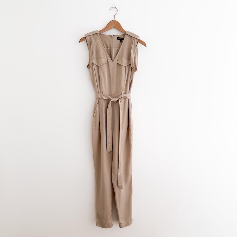 May include: A sleeveless tan jumpsuit with a V-neck, two small chest pockets, and a matching fabric belt. The jumpsuit is hanging on a wooden hanger against a white background. The garment has a zipper closure and shoulder epaulettes.
