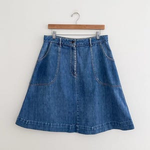 May include: A-line denim skirt in a medium blue wash. The skirt features two large front pockets, a button closure, and a waistband with belt loops. The skirt is hanging on a wooden hanger against a white background.