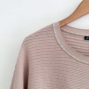 May include: Close-up of a light pink knit sweater with a crew neck and a subtle open weave design. The sweater is hanging on a wooden hanger against a white background. The neckline has a contrasting trim.