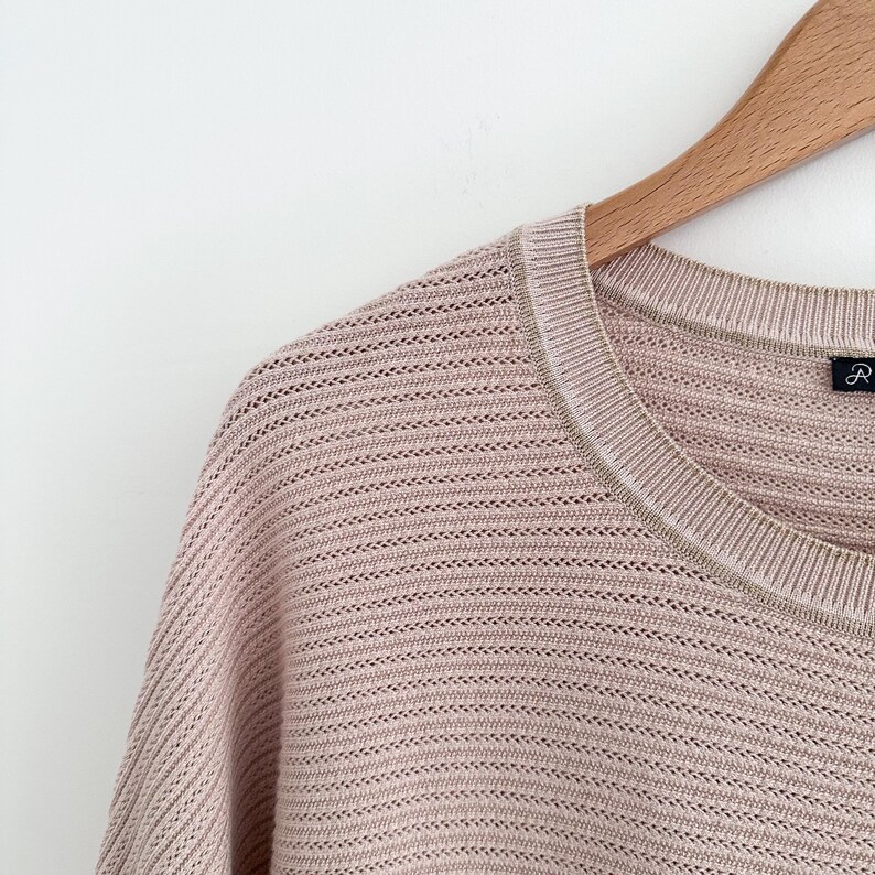 May include: Close-up of a light pink knit sweater with a crew neck and a subtle open weave design. The sweater is hanging on a wooden hanger against a white background. The neckline has a contrasting trim.