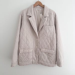May include: A light beige quilted blazer with a classic collar, two front pockets, and two buttons. The jacket is hanging on a wooden hanger against a white background. The jacket has a diamond pattern.