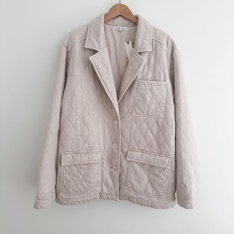 May include: A light beige quilted blazer with a classic collar, two front pockets, and two buttons. The jacket is hanging on a wooden hanger against a white background. The jacket has a diamond pattern.