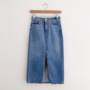 May include: A light blue denim skirt with a front slit, hanging on a wooden hanger. The skirt has a classic five-pocket design and a button and zipper closure. The denim has a faded wash, giving it a vintage look.
