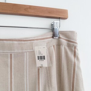 May include: Tan and white striped pants hanging on a wooden hanger. The pants have vertical stripes and a tag attached with product information. The fabric appears to be a linen blend. The pants are size 10.