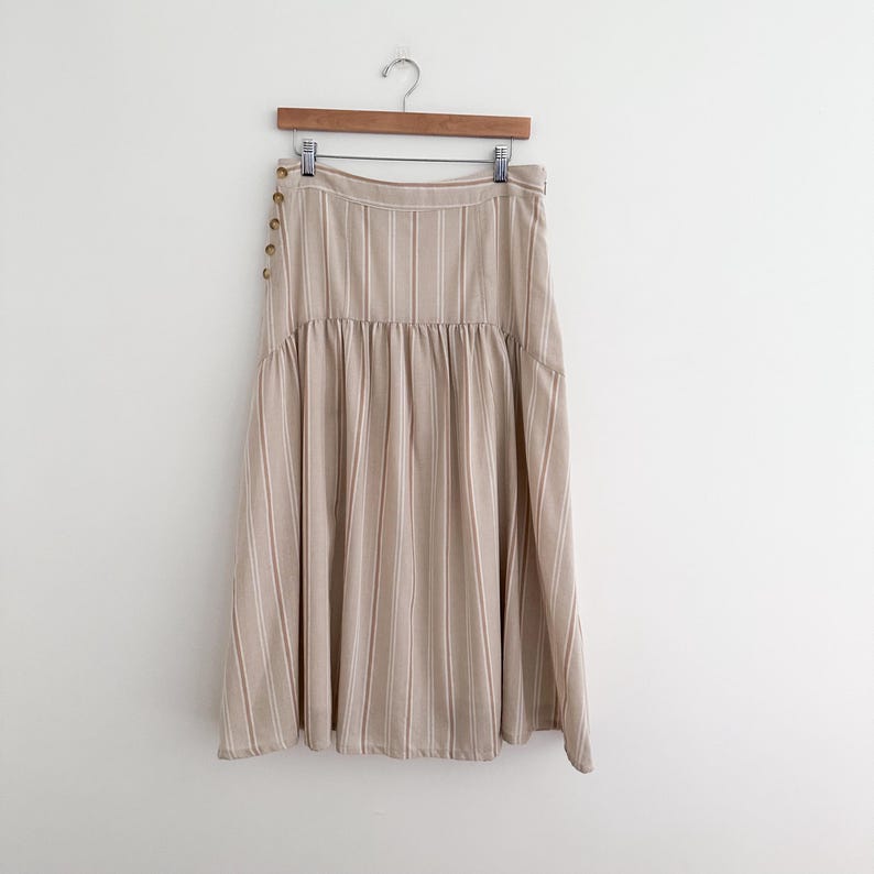 May include: A beige midi skirt with vertical stripes and a gathered waist. The skirt hangs from a wooden hanger and features decorative buttons on the side. The skirt is made of a lightweight fabric.