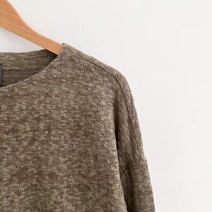 May include: Close-up of a long-sleeved, olive green knit top hanging on a wooden hanger. The fabric has a textured appearance with a subtle, heathered pattern. The top has a crew neck and a relaxed fit, suitable for casual wear.