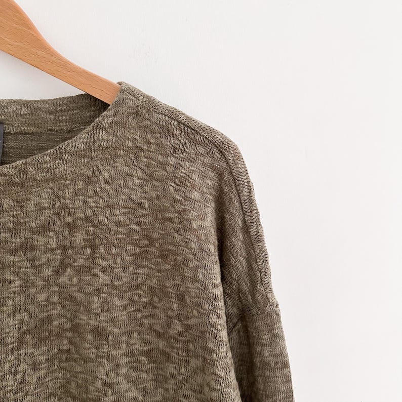 May include: Close-up of a long-sleeved, olive green knit top hanging on a wooden hanger. The fabric has a textured appearance with a subtle, heathered pattern. The top has a crew neck and a relaxed fit, suitable for casual wear.