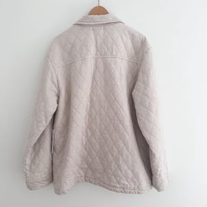 May include: A light beige quilted jacket hanging on a wooden hanger. The jacket has a button-down front, a collar, and diamond-shaped stitching. The jacket is a neutral color and appears to be made of a soft fabric.