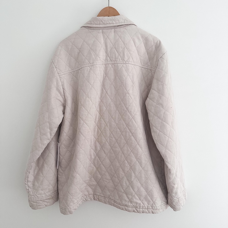 May include: A light beige quilted jacket hanging on a wooden hanger. The jacket has a button-down front, a collar, and diamond-shaped stitching. The jacket is a neutral color and appears to be made of a soft fabric.