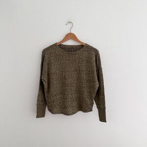 May include: Olive green long-sleeve top with a speckled pattern. The top features a crew neck and solid olive green cuffs. The garment is hanging on a wooden hanger against a white background.