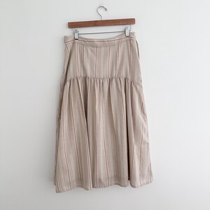 May include: A beige midi skirt with vertical stripes in a lighter beige color. The skirt is hanging on a wooden hanger against a white background. The skirt has a gathered waist and a flared silhouette.