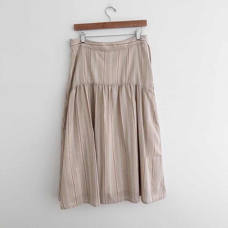 May include: A beige midi skirt with vertical stripes in a lighter beige color. The skirt is hanging on a wooden hanger against a white background. The skirt has a gathered waist and a flared silhouette.