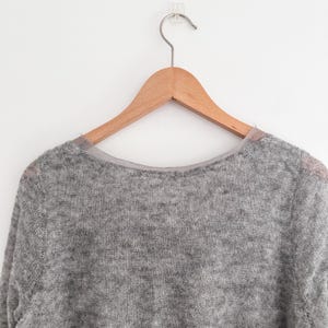 May include: A light gray sweater with a crew neck, hanging on a wooden hanger. The sweater has a soft, textured appearance. The hanger is suspended from a clear adhesive hook on a white wall. The sweater's neckline is trimmed with a sheer, light-colored fabric.