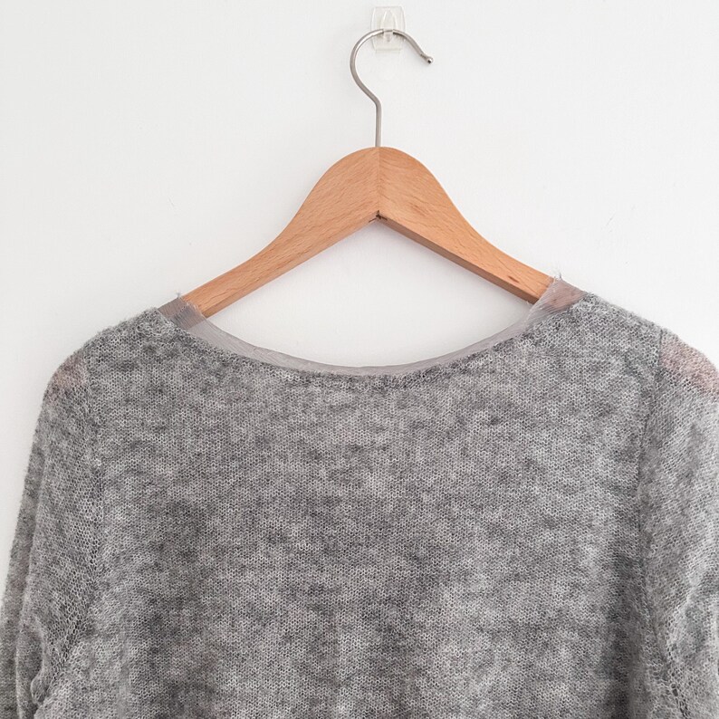 May include: A light gray sweater with a crew neck, hanging on a wooden hanger. The sweater has a soft, textured appearance. The hanger is suspended from a clear adhesive hook on a white wall. The sweater's neckline is trimmed with a sheer, light-colored fabric.