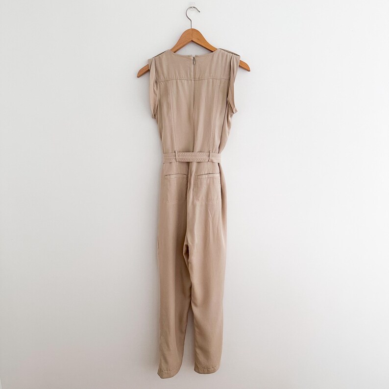 May include: A tan jumpsuit with a sleeveless design, a belted waist, and a zipper closure. The garment is hanging on a wooden hanger against a white wall. The jumpsuit has a tailored fit and appears to be made of a lightweight fabric.