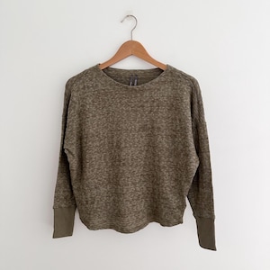 May include: Olive green long-sleeve sweater with a crew neck and ribbed cuffs. The sweater has a textured knit pattern and a relaxed fit. The garment is hanging on a wooden hanger against a white background.