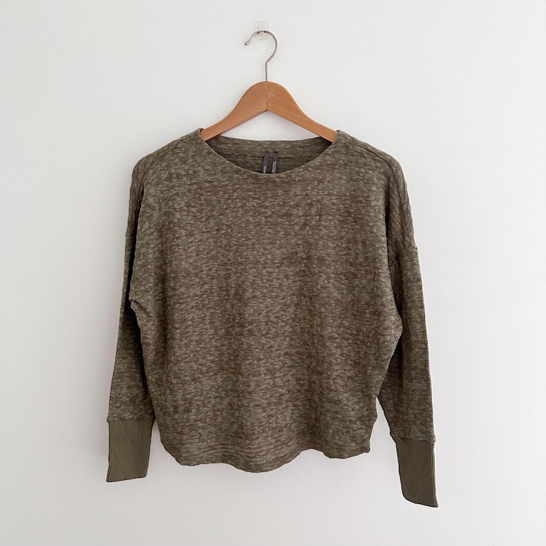 May include: Olive green long-sleeve sweater with a crew neck and ribbed cuffs. The sweater has a textured knit pattern and a relaxed fit. The garment is hanging on a wooden hanger against a white background.