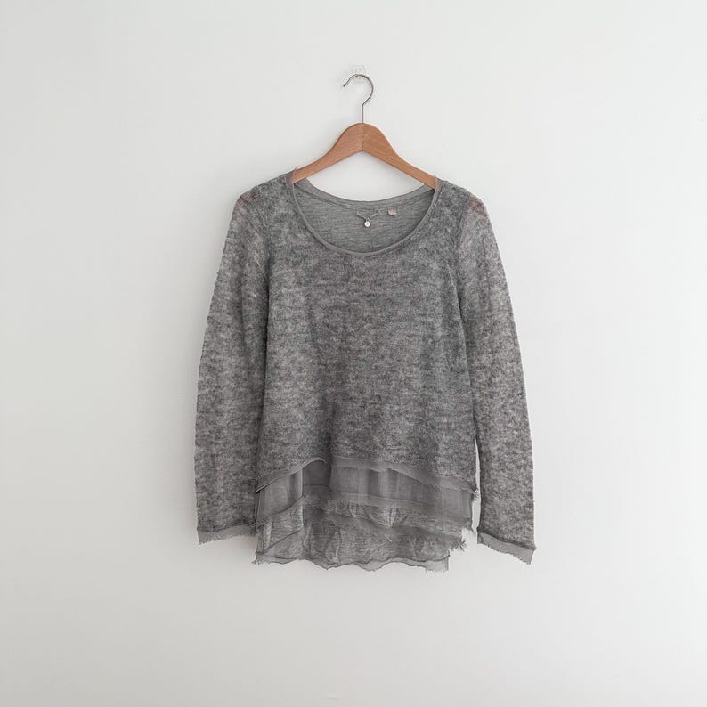 May include: A gray long-sleeved sweater with a scoop neck, hanging on a wooden hanger. The sweater has a layered design with a sheer, ruffled hem. The fabric appears to be a lightweight knit.