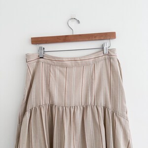 May include: A beige and white striped skirt hanging on a wooden hanger. The skirt has vertical stripes and a ruffled hem. The fabric appears to be a lightweight material, suitable for warmer weather. The skirt is displayed against a plain white background.