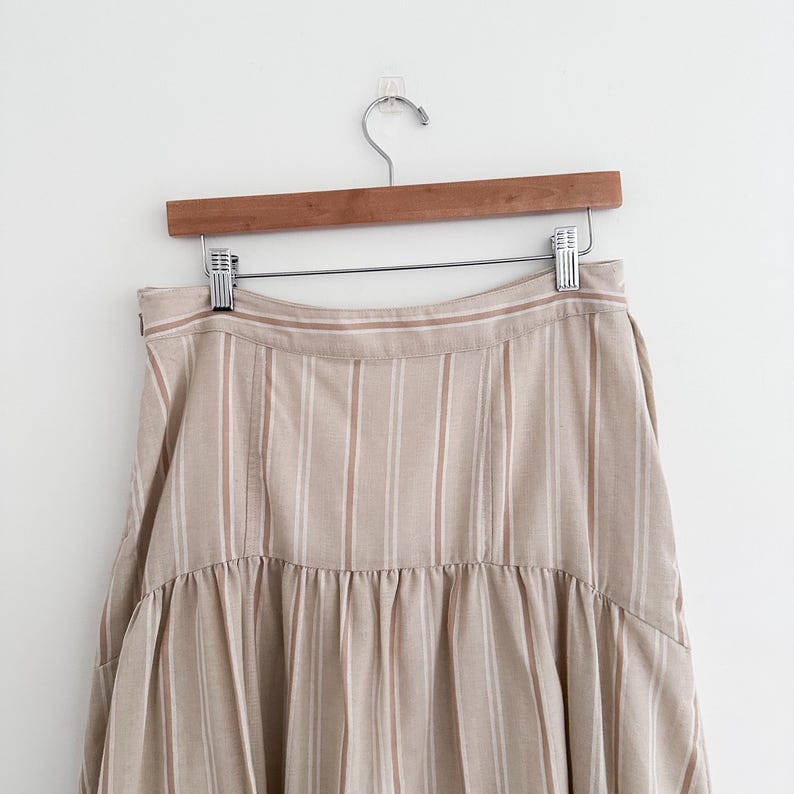 May include: A beige and white striped skirt hanging on a wooden hanger. The skirt has vertical stripes and a ruffled hem. The fabric appears to be a lightweight material, suitable for warmer weather. The skirt is displayed against a plain white background.