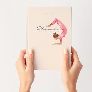 Puede incluir: Una agenda beige con la palabra "Planner" en escritura cursiva y una ilustraci&oacute;n en acuarela de una persona en una postura de yoga. La agenda est&aacute; sujeta por dos manos sobre un fondo blanco.