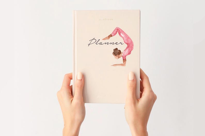Puede incluir: Una agenda beige con la palabra "Planner" en escritura cursiva y una ilustraci&oacute;n en acuarela de una persona en una postura de yoga. La agenda est&aacute; sujeta por dos manos sobre un fondo blanco.