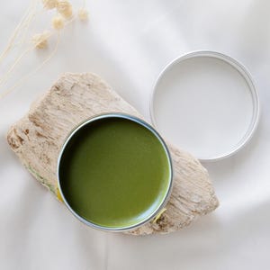 May include: A round metal tin containing a smooth, dark olive green balm, positioned next to its detached silver lid. The container rests upon a piece of light-coloured, textured driftwood, with a small sprig of dried flowers visible nearby.