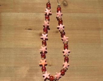 Peut inclure: Un collier orné de perles roses en forme d'étoiles de mer, alternant avec des perles rouges et nacrées. Le collier est doté d'une chaîne dorée et d'un petit pendentif en forme de coquillage. Le bijou est présenté sur une surface en bois.