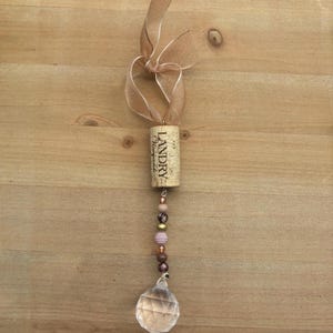 Puede incluir: Adorno decorativo de corcho de vino con una bola de cristal transparente en la parte inferior. El corcho es de Landry Vineyards y está unido a una cadena de cuentas en tonos rosa, marrón y dorado. Una cinta transparente de color marrón claro está atada en la parte superior.