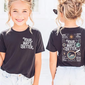 Op de afbeelding: Zwart T-shirt met de tekst "Your Way's Better" in het wit op de voor- en achterkant. De achterkant van het shirt heeft extra tekst en afbeeldingen, waaronder een kruis, een zon en een wereldbol. Het shirt wordt gedragen met een spijkerbroekshort.