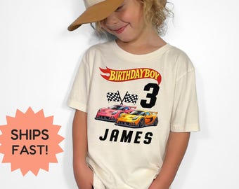 Puede incluir: Una camiseta color crema con un diseño de Hot Wheels para un cumpleaños. La camiseta tiene el texto "BIRTHAY BOY 3 JAMES" con ilustraciones de coches de carreras y banderas a cuadros. El niño lleva una gorra de béisbol gris y marrón.