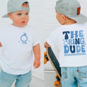 May include: White t-shirt with a blue ring graphic and the name "TOM". The back of the shirt says "THE RING DUDE" in blue lettering. The child is wearing light blue jeans and a gray baseball cap.