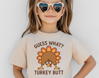 Puede incluir: Una niña lleva gafas de sol en forma de corazón y una camiseta beige. La camiseta presenta un dibujo de un pavo de dibujos animados con el texto "GUESS WHAT? TURKEY BUTT" en naranja y marrón. La niña sonríe y tiene las manos en los bolsillos.