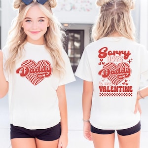 May include: White t-shirt with a red heart-shaped graphic that says "Daddy" in a cursive font. The heart has a red and white checkered pattern. The back of the shirt says "Sorry Boys, Daddy is my Valentine" in red.