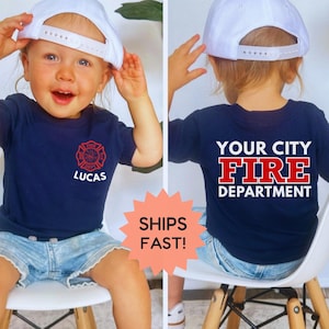 May include: A navy blue t-shirt with a red fire department emblem and the name "Lucas" printed on the front. The back of the shirt displays "YOUR CITY FIRE DEPARTMENT" in white. The child is wearing a white baseball cap and denim shorts.