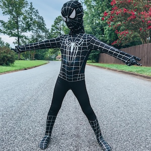 May include: A black and white Spider-Man costume with a hood, featuring a web design and a spider emblem on the chest. The full-body suit has a grid pattern and is worn by a person standing on a road.