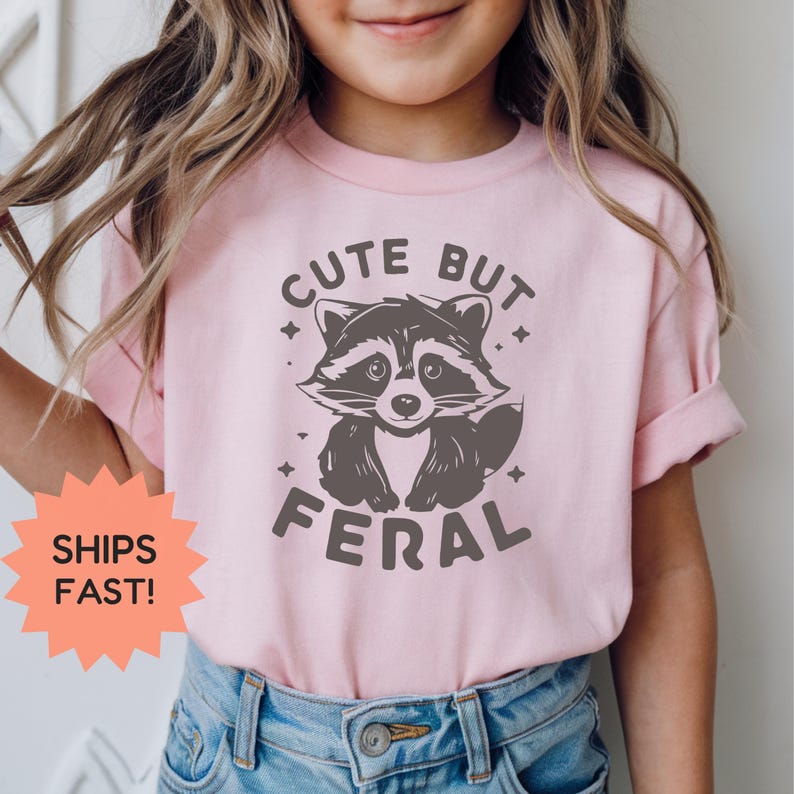 K&ouml;nnte beinhalten: Hellrosa T-Shirt mit einer dunkelgrauen Waschb&auml;r-Grafik und dem Text "CUTE BUT FERAL". Der Waschb&auml;r hat ein freundliches Gesicht. Das T-Shirt wird von einer Person getragen, die eine blaue Jeans tr&auml;gt.