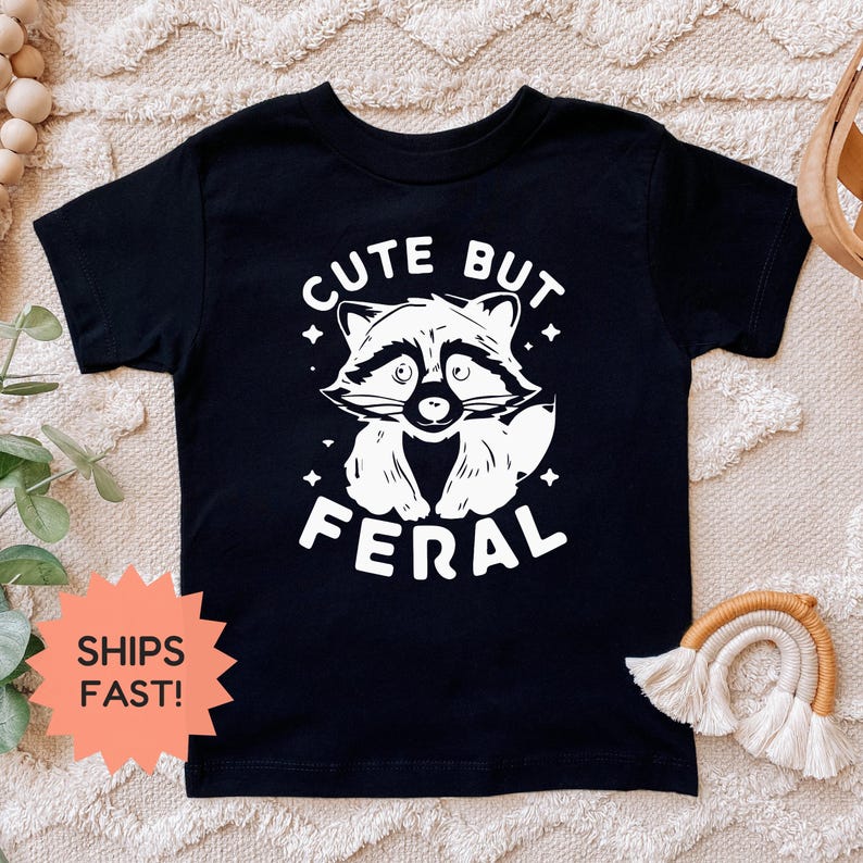 K&ouml;nnte beinhalten: Schwarzes T-Shirt mit wei&szlig;er Grafik eines Waschb&auml;ren und dem Text "CUTE BUT FERAL". Der Waschb&auml;r hat ein detailliertes Gesicht und ist von Sternen umgeben. Kinderbekleidung.